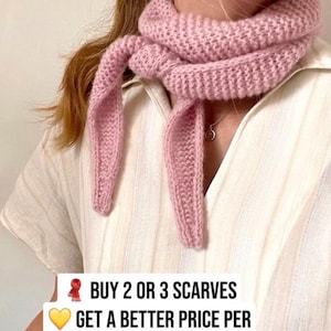 Op de afbeelding: Een lichtroze gebreide sjaal, om de nek geknoopt. De sjaal is gemaakt van dik, getextureerd garen en heeft lange, bungelende uiteinden. De tekst op de afbeelding luidt: "KOOP 2 OF 3 SJAALS KRIJG EEN BETERE PRIJS PER SJAAL".