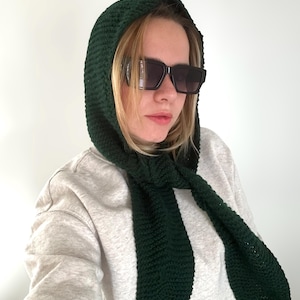 May include: A dark forest green crocheted hood with long scarf-like ends that drape down the front. The hood features a textured, dense stitch pattern and is worn over a light heather gray long-sleeved sweatshirt and black rectangular sunglasses.