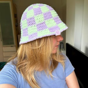 May include: A crochet bucket hat featuring a checkered pattern in alternating light purple and pale green yarn. The hat has a small white rectangular fabric tag attached to the brim with the text MADE WITH LOVE printed in black ink.