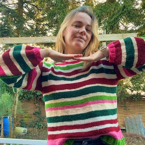 Op de afbeelding: Een kleurrijke gestreepte trui met een ronde hals en lange mouwen. De trui heeft horizontale strepen in rood, groen, roze en wit. De mouwen zijn donkerrood met roze manchetten en de zoom is groen.