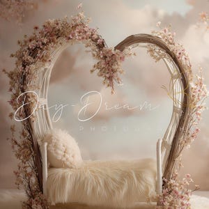 May include: A heart-shaped photography prop with a white woven frame, adorned with pink flowers and branches. It features a fluffy, cream-colored bed and pillow, set on a soft, white rug. The background is a soft, blurred peach and cream color.