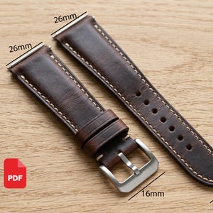 May include: A dark brown leather watch strap featuring white contrast stitching, a silver-toned metal buckle, and two leather keepers. The strap width is labeled as 26mm at the lug end and 16mm at the buckle end, with visible text markings.