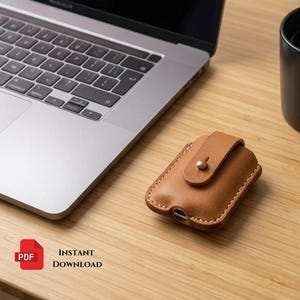 以下が含まれることがあります： ワイヤレスイヤホン用のタンカラーのレザー製保護ケース。フラップと金属製のスタッドボタンが付いています。ケースの縁にはステッチが施され、底面には充電用の切り込みがあります。ノートパソコンと「HUSTEBYTRADE」と書かれた黒いマグカップが近くに置かれています。