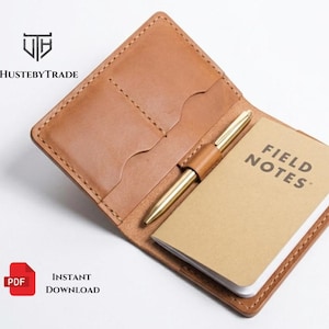 May include: A tan leather notebook cover with a small notepad and a gold pen. The notepad is labeled "FIELD NOTES". The cover has two card slots and a pen holder. The item is open and displayed on a white surface.