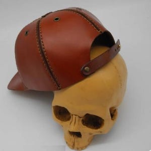 K&ouml;nnte beinhalten: Eine braune Baseballkappe aus Leder mit sichtbaren N&auml;hten, Metall&ouml;sen zur Bel&uuml;ftung und einem verstellbaren Lederriemen mit Metalldruckknopf auf der R&uuml;ckseite, pr&auml;sentiert auf einem beigen Modell eines menschlichen Sch&auml;dels.