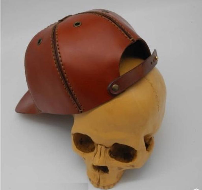K&ouml;nnte beinhalten: Eine braune Baseballkappe aus Leder mit sichtbaren N&auml;hten, Metall&ouml;sen zur Bel&uuml;ftung und einem verstellbaren Lederriemen mit Metalldruckknopf auf der R&uuml;ckseite, pr&auml;sentiert auf einem beigen Modell eines menschlichen Sch&auml;dels.