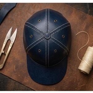 K&ouml;nnte beinhalten: Eine marineblaue Baseballkappe aus Leder mit sechs Paneelen, wei&szlig;en Kontrastn&auml;hten, einem zentralen Knopf und sechs metallischen &Ouml;sen. Die Kappe wird zusammen mit einer Messing-Fadenschere und einer Garnrolle aus hellem Faden pr&auml;sentiert.