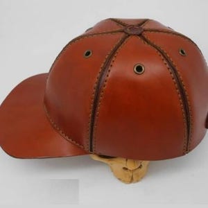 K&ouml;nnte beinhalten: Eine braune Baseballkappe aus Leder mit sechs Paneelen, die durch sichtbare N&auml;hte verbunden sind. Die Kappe verf&uuml;gt &uuml;ber metallische &Ouml;sen zur Bel&uuml;ftung auf jedem Paneel, einen gebogenen Schirm und einen zentralen Knopf an der Oberseite.
