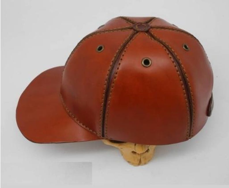 K&ouml;nnte beinhalten: Eine braune Baseballkappe aus Leder mit sechs Paneelen, die durch sichtbare N&auml;hte verbunden sind. Die Kappe verf&uuml;gt &uuml;ber metallische &Ouml;sen zur Bel&uuml;ftung auf jedem Paneel, einen gebogenen Schirm und einen zentralen Knopf an der Oberseite.