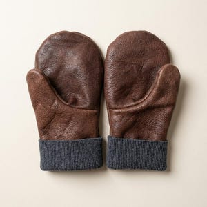 May include: A pair of brown leather mittens featuring a textured, pebbled surface and dark gray ribbed knit cuffs. The mittens are shaped with a distinct thumb section and a rounded top, showcasing the natural grain of the leather material.