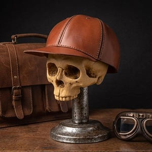 K&ouml;nnte beinhalten: Eine braune Baseballkappe aus Leder auf einem menschlichen Sch&auml;delmodell, das auf einem Metallst&auml;nder montiert ist. Neben dem Sch&auml;del befinden sich eine braune Ledertasche mit Schnalle und eine dunkel get&ouml;nte Fliegerbrille mit braunem Rahmen.