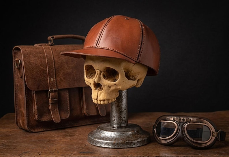 K&ouml;nnte beinhalten: Eine braune Baseballkappe aus Leder auf einem menschlichen Sch&auml;delmodell, das auf einem Metallst&auml;nder montiert ist. Neben dem Sch&auml;del befinden sich eine braune Ledertasche mit Schnalle und eine dunkel get&ouml;nte Fliegerbrille mit braunem Rahmen.