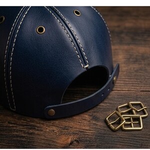 K&ouml;nnte beinhalten: Eine dunkelblaue Baseballkappe aus Leder mit wei&szlig;en Kontrastn&auml;hten, messingfarbenen Metall&ouml;sen zur Bel&uuml;ftung und einem verstellbaren Lederriemen mit messingfarbenem Druckknopfverschluss, begleitet von drei kleinen messingfarbenen Metallschnallen.