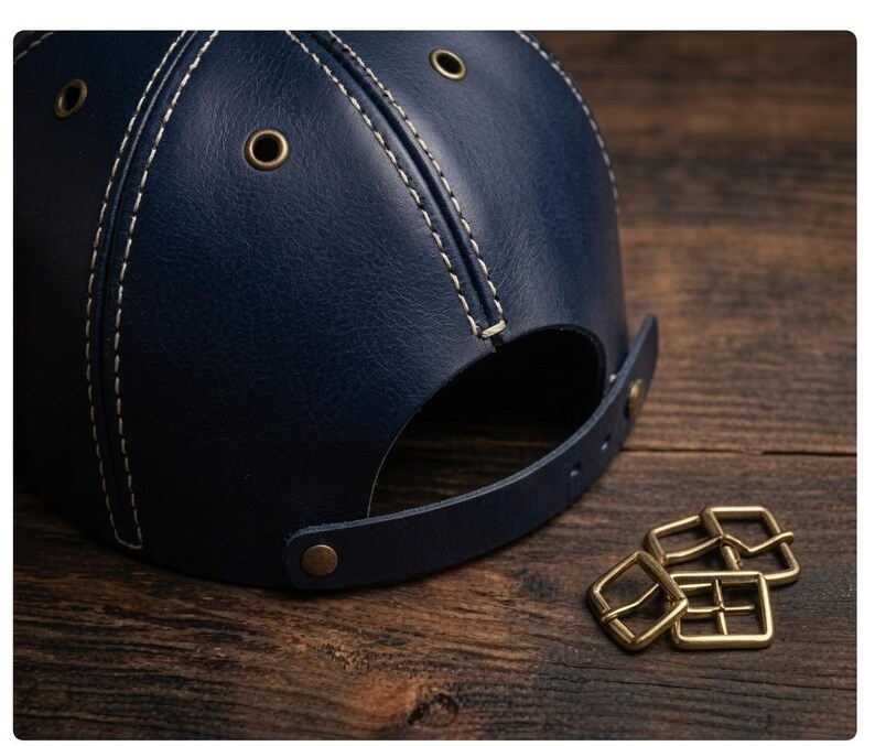 K&ouml;nnte beinhalten: Eine dunkelblaue Baseballkappe aus Leder mit wei&szlig;en Kontrastn&auml;hten, messingfarbenen Metall&ouml;sen zur Bel&uuml;ftung und einem verstellbaren Lederriemen mit messingfarbenem Druckknopfverschluss, begleitet von drei kleinen messingfarbenen Metallschnallen.