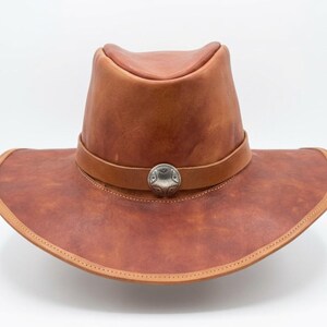 May include: A brown leather cowboy hat featuring a wide, curved brim and a pinched crown. A matching leather band encircles the base of the crown, secured at the front by a circular, silver-toned metal concho with intricate embossed detailing.