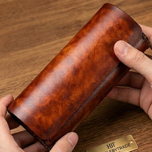 May include: A cylindrical, brown leather case with a rich, textured finish. The case is held by two hands, showcasing its polished surface and the intricate details of the leatherwork. A small, gold-colored tag is visible.