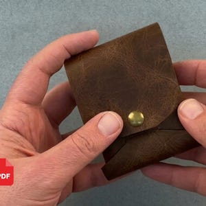 May include: A small, square-shaped brown leather pouch featuring a distressed texture and a single brass-colored metal snap button closure centered on the front flap. The pouch is constructed from a single piece of folded leather with visible stitching.