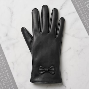 Könnte beinhalten: Ein schwarzer Lederhandschuh mit einer kleinen, dekorativen Schleife am Handgelenk. Der Handschuh liegt auf einer weißen Marmoroberfläche neben einer grauen Schneidematte mit sichtbaren Maßangaben in Zentimetern.