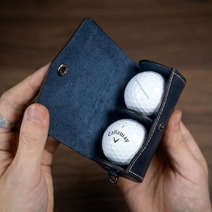 Puede incluir: Un estuche de cuero azul oscuro para pelotas de golf con cierre de broche, que contiene dos pelotas de golf blancas. Una pelota presenta el logotipo y el texto Callaway en negro, mientras que la otra muestra texto negro. El estuche tiene costuras visibles.