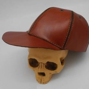 K&ouml;nnte beinhalten: Eine strukturierte Baseballkappe aus glattem, poliertem braunem Leder. Die Kappe verf&uuml;gt &uuml;ber sichtbare N&auml;hte, die die Paneele verbinden, sowie kleine metallische &Ouml;sen an den Seiten zur Bel&uuml;ftung und ruht auf einem beigen menschlichen Sch&auml;delmodell.
