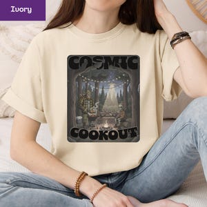 May include: Ivory-colored t-shirt featuring a graphic of a cosmic cookout scene. The design includes a Bigfoot and an alien grilling food around a campfire, with the words "COSMIC COOKOUT" in a black frame. The shirt is a casual wear item.