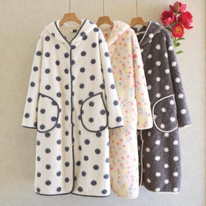 Puede incluir: Tres batas de forro polar con capucha colgando de perchas de madera. Las batas son de color crema con lunares azul marino, crema con estampado floral y gris con lunares blancos. Cada bata tiene bolsillos y cierre de botón.