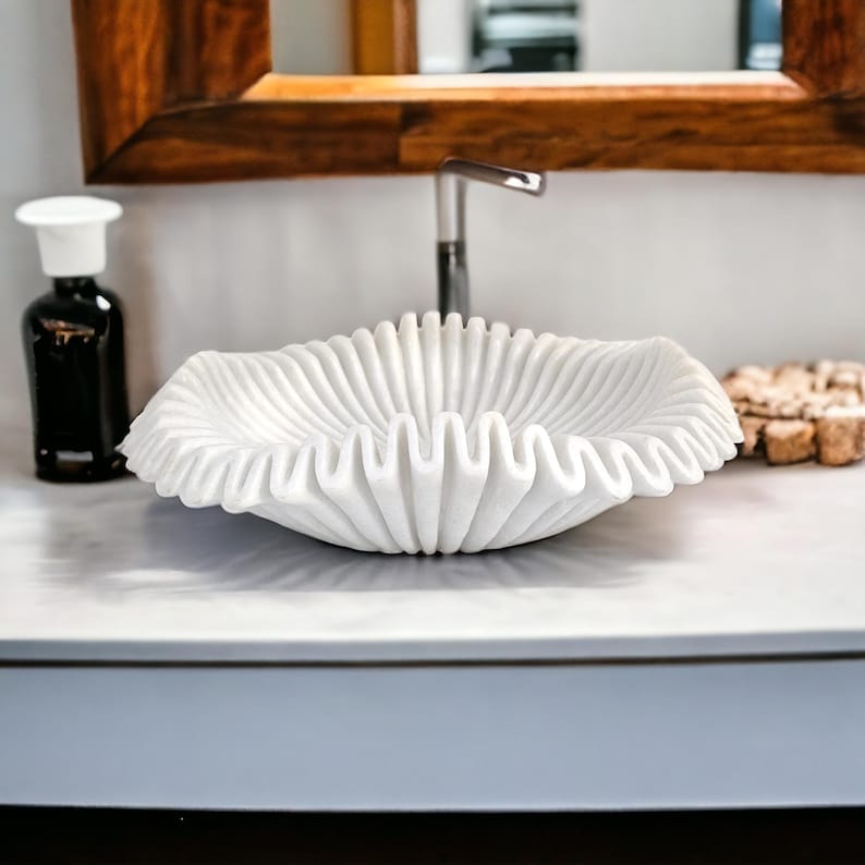 May include: A white marble sink with a wavy, textured design. The sink is in the shape of a seashell and sits on a white countertop. A black bottle with a white top is to the left of the sink.