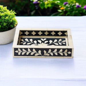 May include: Rectangular decorative tray with a black and white leaf design. The tray has a cream-colored frame with black accents and a black interior. The tray is made of a durable material and is suitable for home decor.