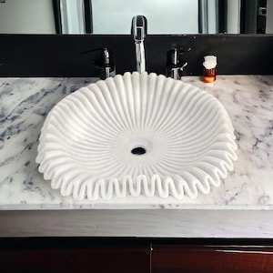 May include: A white marble sink with a unique, textured design. The sink has a circular shape with a ruffled edge and radial grooves, creating a decorative and functional bathroom fixture.