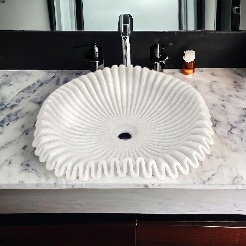 May include: A white marble sink with a unique, textured design. The sink has a circular shape with a ruffled edge and radial grooves, creating a decorative and functional bathroom fixture.