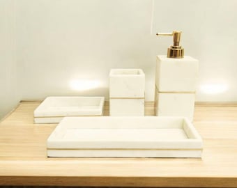 May include: A set of white marble bathroom accessories with gold accents. The set includes a soap dispenser, a toothbrush holder, and two trays. The items are arranged on a light wood surface.
