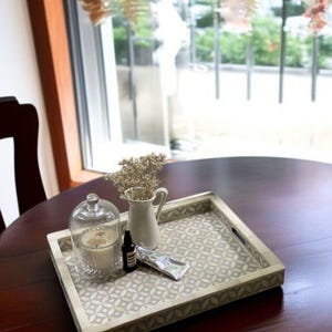 May include: A decorative square tray with a geometric pattern holds a glass cloche with a candle, a small white pitcher with dried flowers, a dark bottle, and a tube of cream. The tray is on a dark wooden table near a window.