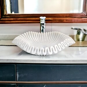 May include: A white marble vessel sink with a unique ruffled edge design. The sink has a fluted interior and is set on a marble countertop. A chrome faucet and mirror are visible in the background, creating a luxurious bathroom aesthetic.