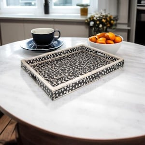 May include: A rectangular decorative tray with a black and white patterned design. The tray has a light-colored frame and handles. A white bowl of oranges, a blue teacup and saucer, and flowers are on a marble table.