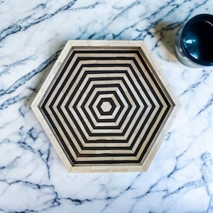 May include: Hexagonal decorative tray with a black and white geometric pattern. The tray has a light-coloured frame and a black and white striped interior design. A dark-coloured cup is visible in the upper right corner. The tray is on a marble surface.