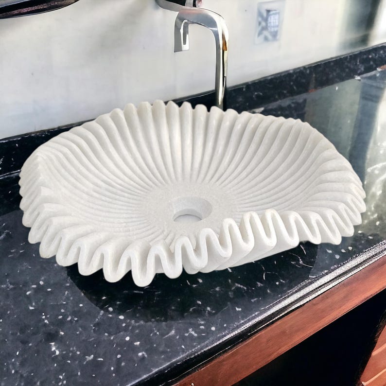 May include: A white marble sink with a unique, ruffled design. The sink has a central drain and is set against a black countertop. The faucet is chrome.