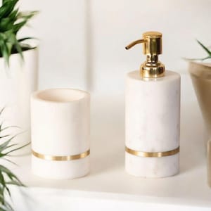 Op de afbeelding: Een set van een witte marmeren zeepdispenser en beker. De dispenser heeft een gouden pomp en een gouden band rond de basis. De beker heeft een gouden band rond de basis. De set staat op een wit oppervlak.