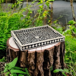 May include: A rectangular decorative tray with a black and white floral design. The tray is made of wood and has a raised edge. The tray is sitting on a tree stump in a natural outdoor setting.