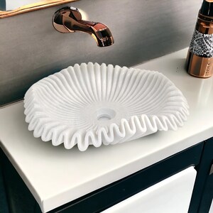 May include: A white marble sink with a ruffled edge design. The sink is round with a central drain hole and sits on a white countertop. A copper faucet is mounted on the wall above the sink.