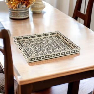 May include: A square decorative tray with a black and cream floral design. The tray has a rectangular shape with a black border and handles. The interior features a detailed floral pattern. The tray is placed on a light brown wooden table.