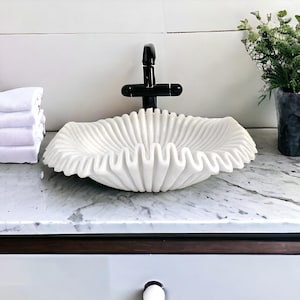 May include: A white, ruffled marble sink basin with a black faucet. The sink has a unique, wave-like design. A stack of white towels is on the left. The sink is on a marble countertop.