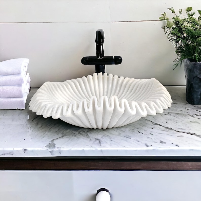 May include: A white, ruffled marble sink basin with a black faucet. The sink has a unique, wave-like design. A stack of white towels is on the left. The sink is on a marble countertop.