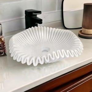 May include: A white, intricately carved marble sink with a ruffled edge. The sink is round with a central drain hole and sits on a white countertop. A black faucet is visible in the background, along with a wooden cup.