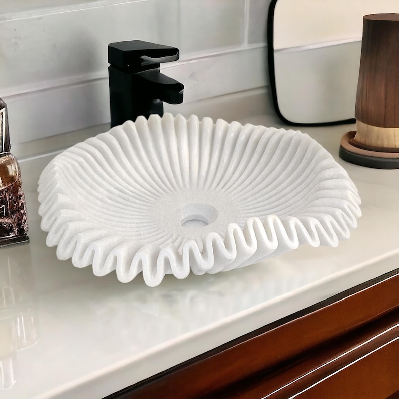 May include: A white, intricately carved marble sink with a ruffled edge. The sink is round with a central drain hole and sits on a white countertop. A black faucet is visible in the background, along with a wooden cup.