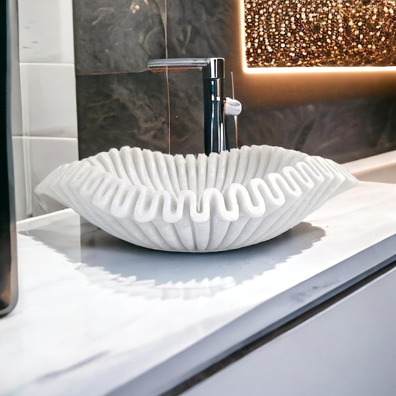 May include: A white marble sink with a ruffled edge design. The sink is in a bathroom setting with a chrome faucet and a decorative light fixture in the background. The sink's wavy design adds a unique touch to the bathroom decor.