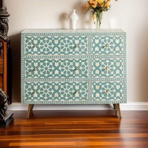 Puede incluir: Cómoda de seis cajones con un patrón geométrico turquesa y blanco. La cómoda tiene herrajes y patas doradas. Un jarrón con flores y una pequeña estatua están encima. La cómoda está sobre un suelo de madera.
