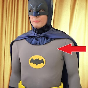 Könnte beinhalten: Ein grauer Batman-Anzug mit einem gelben Fledermaus-Emblem auf der Brust. Das Kostüm umfasst einen dunkelblauen Umhang, eine Kapuze und eine Maske. Ein gelber Werkzeuggürtel ist ebenfalls zu sehen.