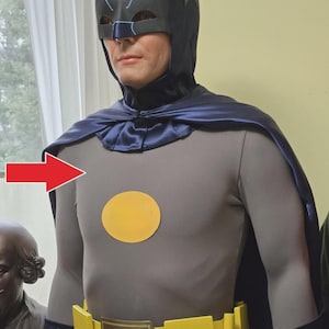 Könnte beinhalten: Ein Ganzkörperkostüm mit einer dunkelblauen Maske mit spitzen Ohren, einem grauen Langarmshirt mit einem gelben Kreis auf der Brust, einem dunkelblauen Satin-Umhang und einem leuchtend gelben Gürtel mit rechteckiger Schnalle und Fledermaus-Emblem.