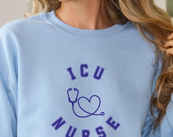 May include: A light blue crewneck sweatshirt featuring dark blue text that reads ICU NURSE in an arched design. Centered between the words is a dark blue line art graphic of a stethoscope shaped into a heart outline on the chest area.