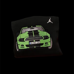 Op de afbeelding: Zwart sweatshirt met een geborduurd ontwerp van een groene sportwagen met zwarte racestrepen. De auto heeft een kentekenplaat met de tekst "AI-EM-256". Een wit Jumpman-logo is rechtsboven geborduurd.